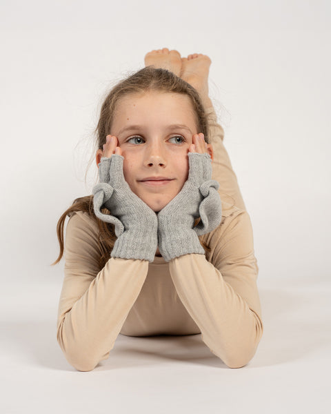 Pige liggende med hovedet hvilende på hænderne, iført lysegrå strikkede konvertible vanter med åbne vanteklapper.