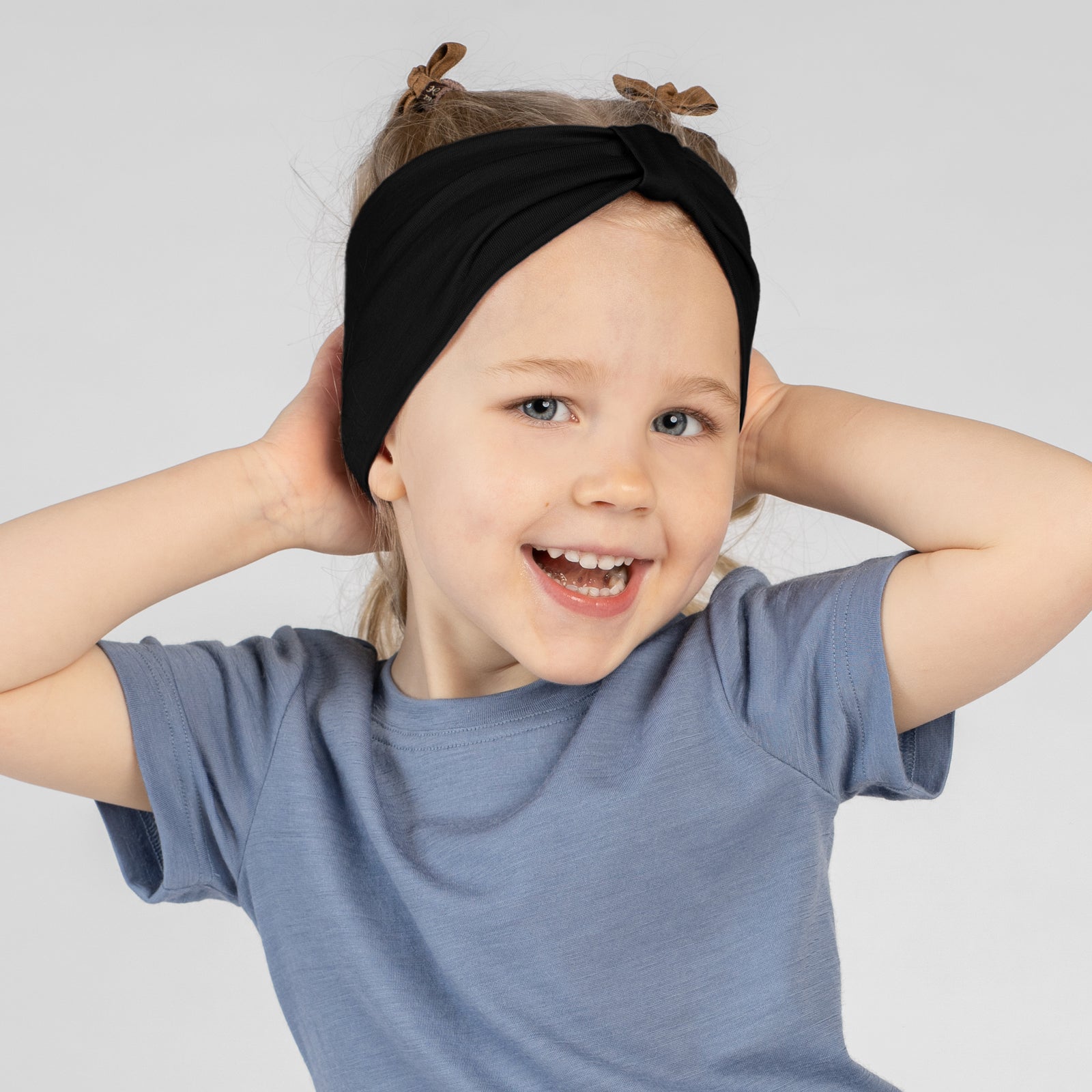 A close-up studio portrait of a young girl smiling directly at the camera. She wears a short-sleeved dusty blue top and a matching dusty blue ribbon headband. Her hands are gently resting behind her neck against a plain light grey background.