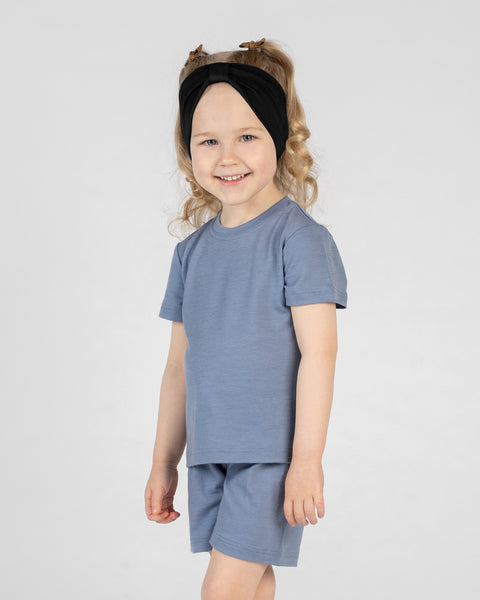 A studio portrait of a young girl standing and smiling directly at the camera against a plain light grey background. She is wearing a matching short-sleeved top and shorts set in blue, along with a black ribbon headband in her hair.