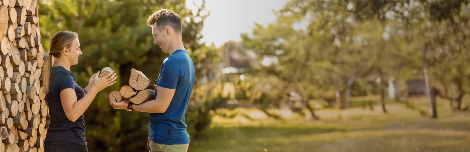 A woman and a man standing outdoors next to a textured tree trunk, with the man holding a small brown dog while they look at each other.