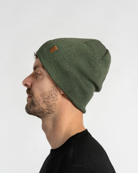 A man with light skin, blue eyes, and a beard. He wears a dark green colored menique knit beanie with a small brown patch on the side, and a black crew-neck shirt.
