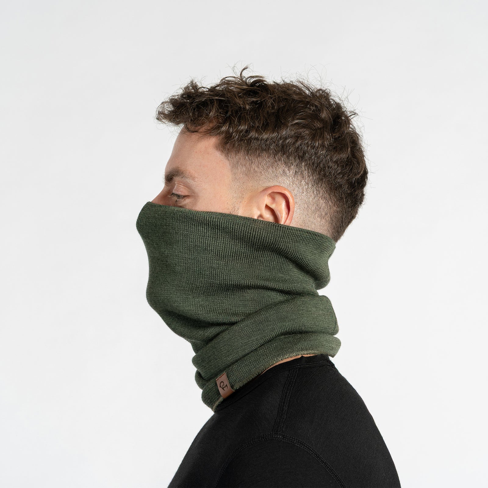 A man with short, dark hair and light eyes looks toward the camera while modeling a dark green knitted neck gaiter. The gaiter is pulled up over his nose and mouth, covering the lower portion of his face, and is set against a plain white background.