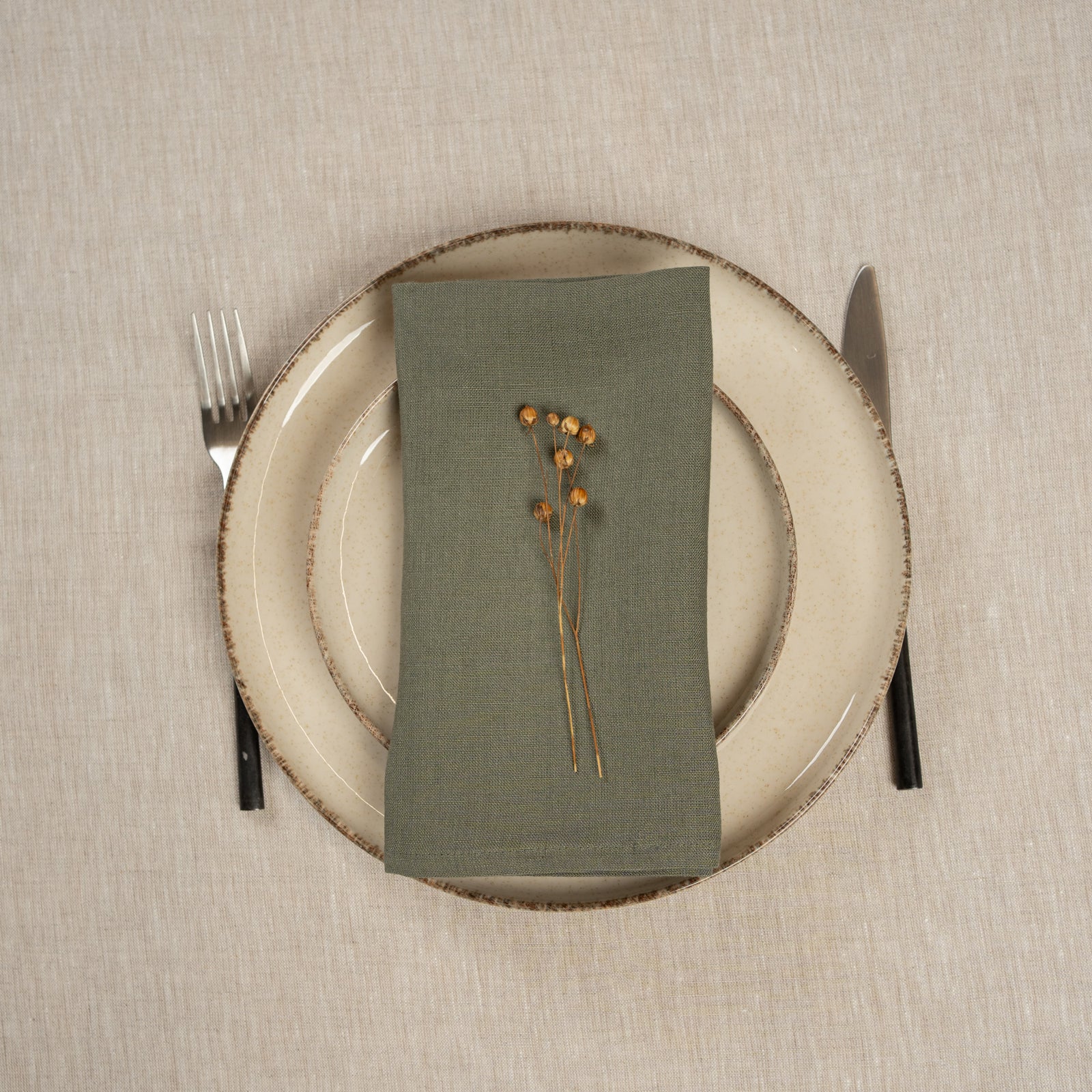 Menique hørserviet i krydret gul farve, lagt oven på to stablede tallerkener. Der ligger bestik ved siden af tallerkenerne. Baggrunden er en dug i naturlig farvet hør.