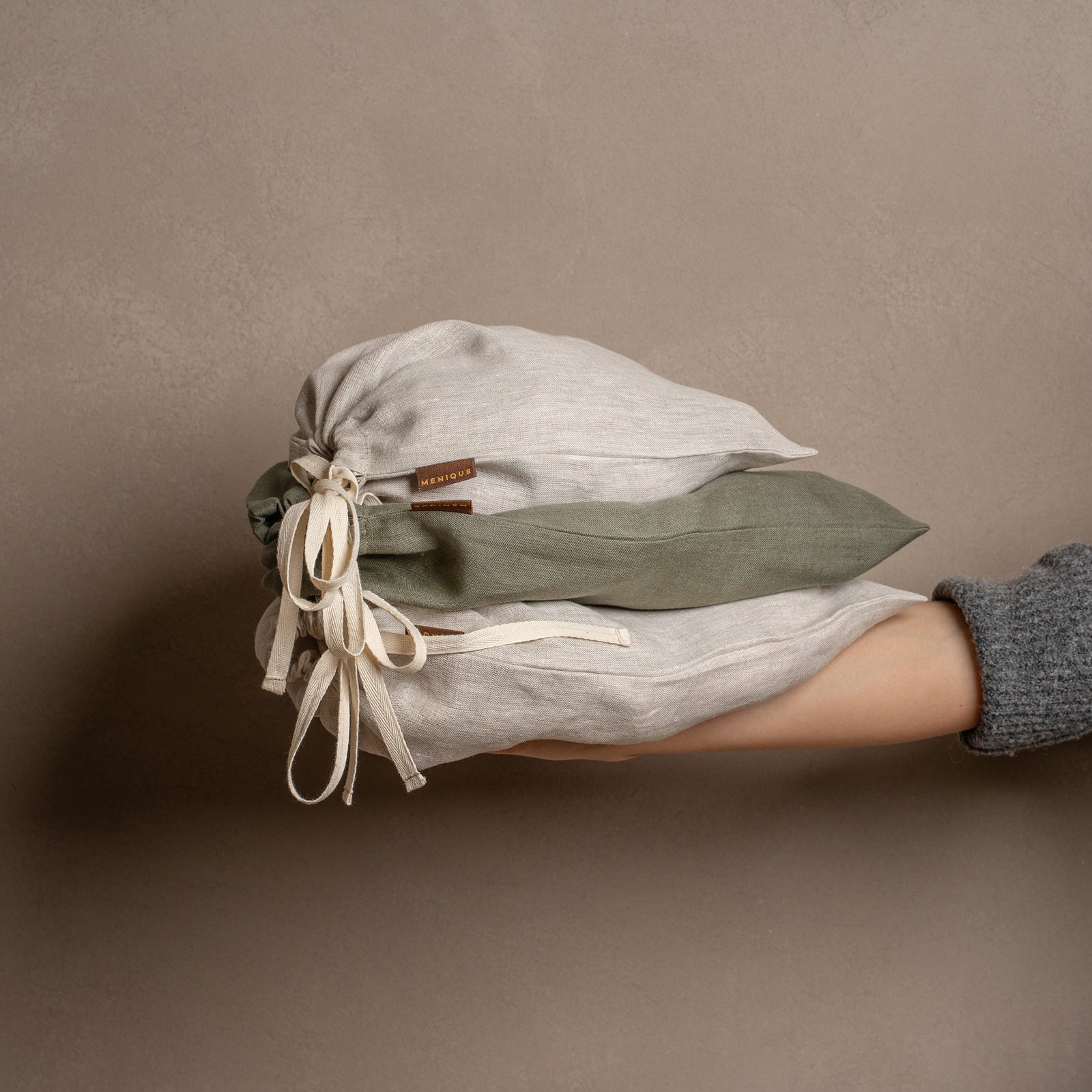 Fem lukkede linnedposer med snøre i forskellige størrelser og farver i natur og stengrøn er arrangeret på et struktureret jutegulvtæppe mod en varm brun væg.