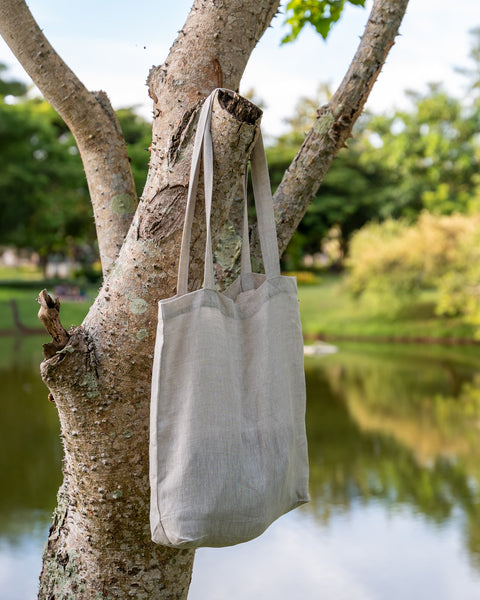 Menique natural color linen tote bag with long handles hangs from a branch of a light-colored tree. In the blurred background, there is a body of water reflecting the sky, surrounded by green grass and trees under a partly cloudy sky.