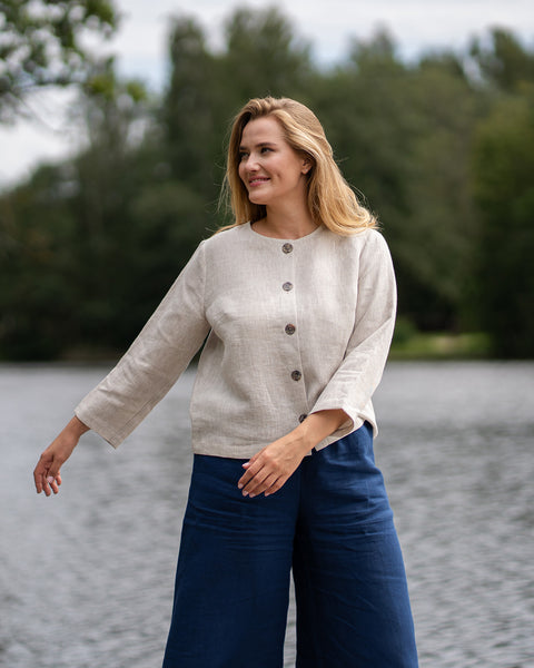 A woman with blonde hair stands near a lake wearing a cream sweater and wide-leg blue trousers. She poses with one arm extended outwards while looking thoughtfully away from the camera.