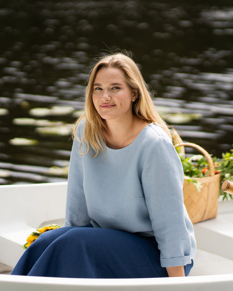 A woman sits calmly in a white rowboat, smiling warmly toward the camera. She wears a light blue top and dark blue pants, with bright sunflowers resting in her lap.
