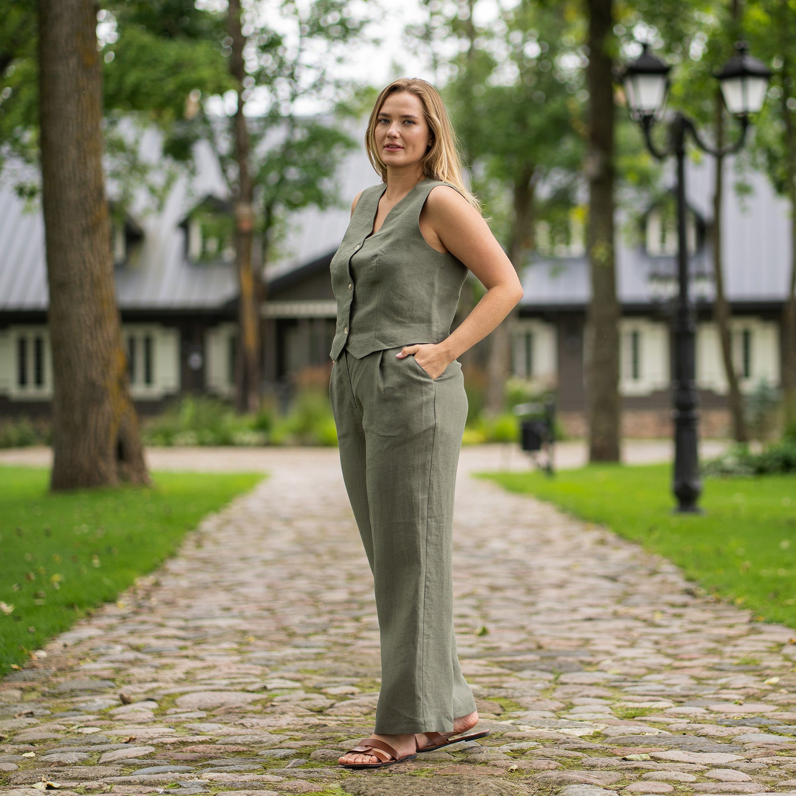 En person ses fra livet og ned iført stengrønne, højtaljede, vidde bukser i hør. De har sorte sandaler med åbne tæer og tynde remme på, og hænderne hviler på hofterne. Omgivelserne er udendørs på en asfaltsti med et sort metalhegn og grønt løv i baggrunden.