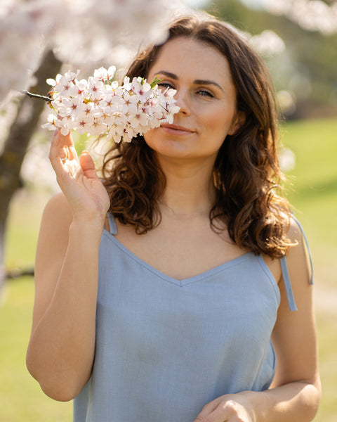 En kvinde iført en skyblå linnednatkjole står ved siden af et træ dækket af hvide blomster og dufter blidt til en klase blomster.