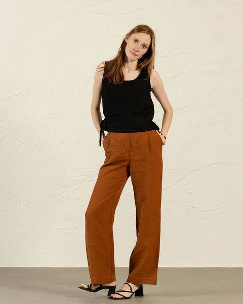 Et studieportræt af en kvinde med langt, lyst brunt hår, der står med hænderne i lommerne og ser direkte ind i kameraet. Hun har en sort ærmeløs top med rund halsudskæring og bindebånd i siderne på, kombineret med rustfarvede bukser med vide ben. Hun har også sorte sandaler med åben tå og blokhæl på. Baggrunden er en ensfarvet, lys væg.