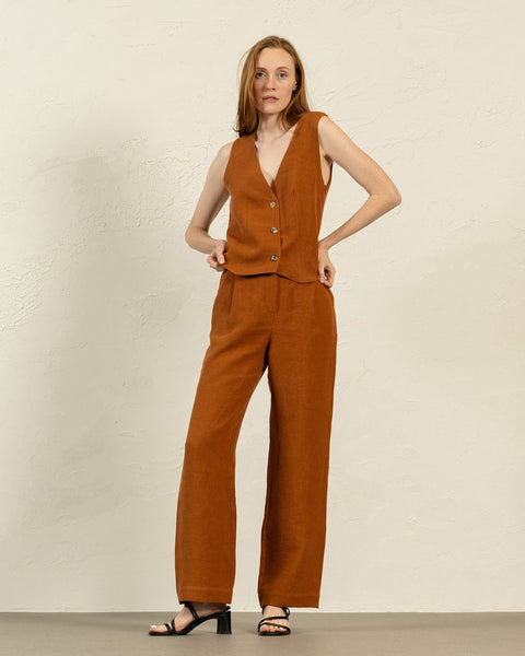 En lys hudet kvinde med skulderlangt brunt hår står vendt fremad og viser en hørvest i mandelbrun og menique bukser med vide ben. Hun har hænderne hvilende på hofterne og bærer sorte sandaler med åben tå og lav hæl. Baggrunden er en struktureret lys beige væg og en lys grå gulv.