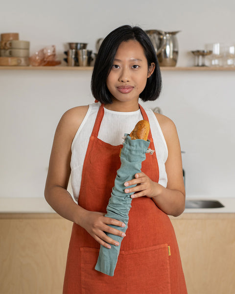 Woman wearing a rust-colored linen bib apron holding bread wrapped in a linen bread bag in a modern kitchen.