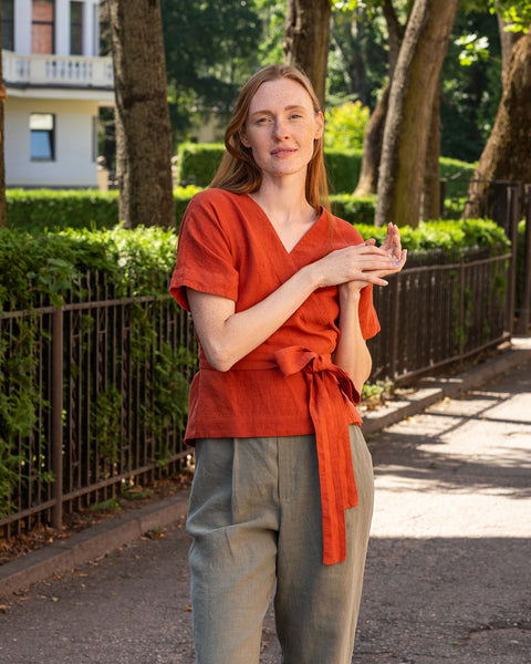 En person med lys hud og mørkt hår står i profil og ser ned mod sine hænder, som er krydsede foran maven. Personen har en hørviklebluse på med korte, løstsiddende ærmer og stengrønne bukser med vide ben. I baggrunden ses et mørkt metalhegn og frodigt grønt løv.