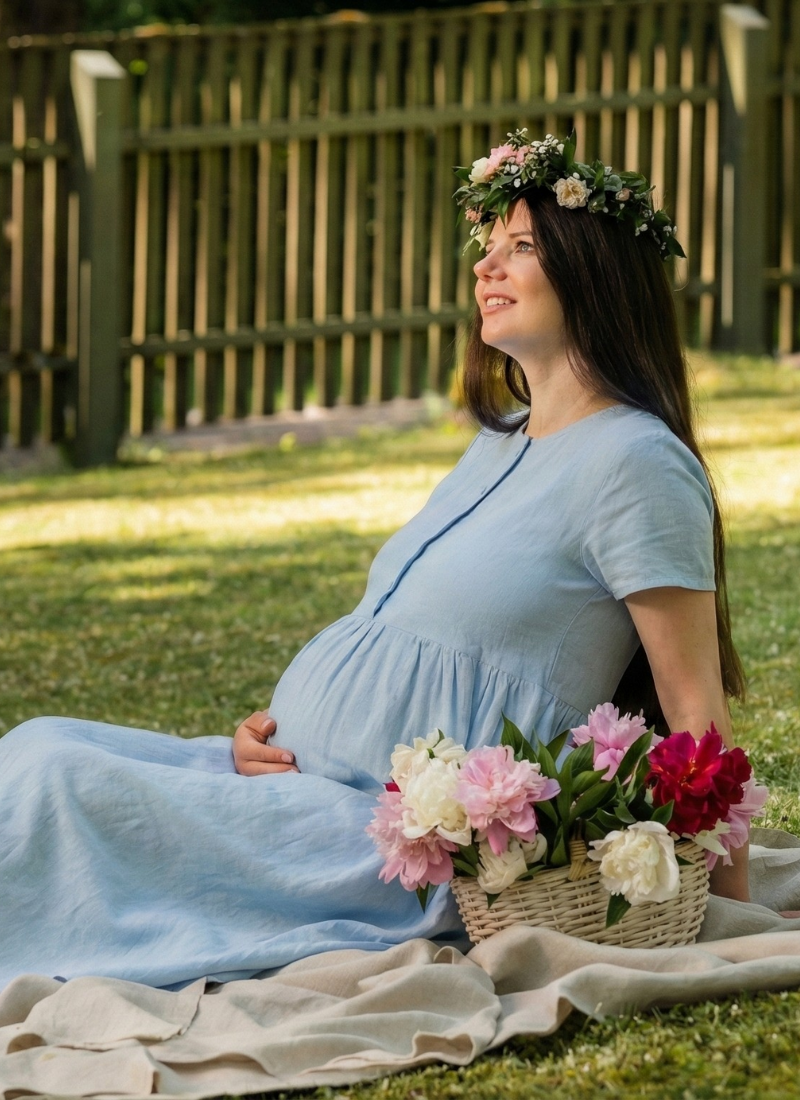 En gravid kvinde sidder på et tæppe i en grøn have iført en lyseblå hørgraviditetskjole og en fin blomsterkrans. Hun hviler blidt sin hånd på sin mave, mens hun kigger opad, med en kurv fyldt med friske blomster ved siden af sig, hvilket skaber en fredfyldt og glædesfyldt sommerscene.