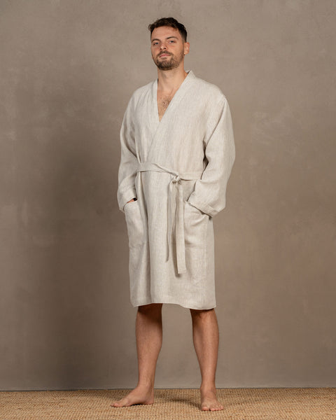 A man, looking at the camera, wearing the menique men's linen bathrobe in natural color, with both of his hands inside the side pockets. The front tie is tied into a loose knot, the robe features a v-neck cut at the front. The bathrobe is knee-length.