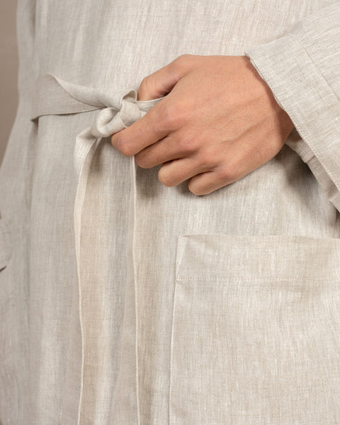 A close-up shot of a hand tying the belt of a natural color linen robe, showing the fabric texture and pocket detail.