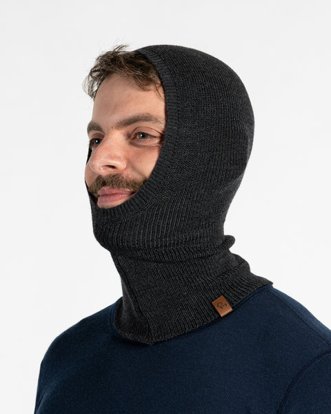 A man with dark hair, a beard, and a mustache is seen from the side, wearing a dark gray knitted balaclava that covers his head and neck, with an opening around his face. He is also wearing a dark blue long-sleeved shirt and appears to be looking off to his right with a slight smile.
