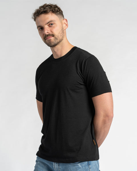 En mand iført en sort t-shirt i merinould og blå jeans ses fra brystet og op mod en lys baggrund. Han har kort brunt hår og et let skæg, og kigger let til siden. Menique-sort t-shirten er i fokus og viser dens pasform og udseende.
