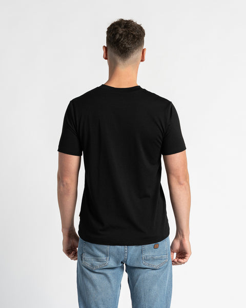 Bagside af en mand iført en sort t-shirt og lyseblå jeans, stående mod en hvid baggrund. Den sorte t-shirt er i fokus og viser dens almindelige pasform og design bagfra. Manden er placeret centralt og viser tydeligt bagsiden af tøjet.