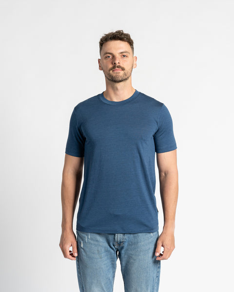 En mand iført en t-shirt i denimfarve af 100 % merinould og lyseblå jeans står midt på en hvid baggrund. Menique-t-shirten er i fokus og viser dens almindelige pasform og udseende. Manden har kort brunt hår og let skæg og ser direkte på beskueren.