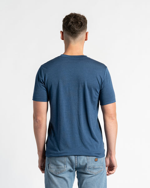 Bagside af en mand iført en menique t-shirt i merinould og lyseblå jeans, stående mod en hvid baggrund. T-shirten i denimfarve er i fokus og viser dens pasform og design bagfra. Manden er placeret centralt og viser tydeligt bagsiden af tøjet.