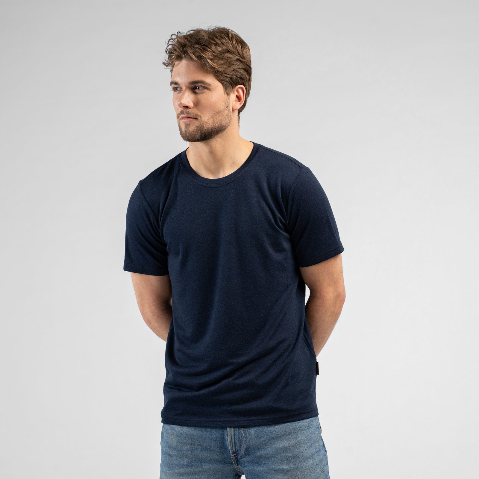 A studio portrait of a man sitting on a black stool against a plain white background. He is leaning slightly forward with his hands resting on his upper thighs, looking directly at the camera. He is wearing a dark navy blue short-sleeved crew-neck t-shirt and blue jeans.