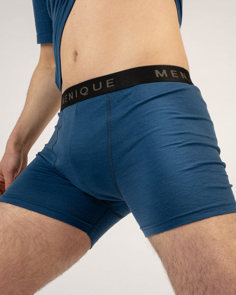 A close-up angled shot of a man's torso and thighs wearing denim blue short boxer briefs with a black "MENIQUE" waistband. He is pulling up a matching denim blue t-shirt to reveal his midriff against a light grey background.