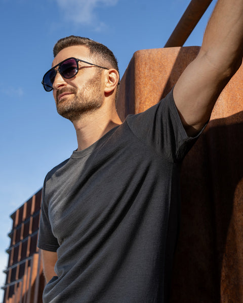 A man wearing a charcoal grey t-shirt and dark sunglasses leans with his left arm stretched out against a large, rusted steel structure. He is looking off to the left under a bright blue sky.