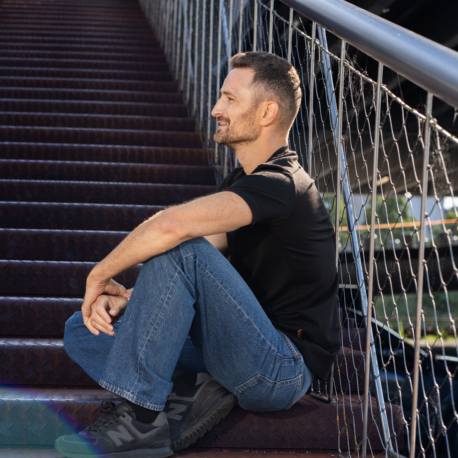 A smiling man sitting on outdoor metal stairs. He is wearing a short-sleeve black V-neck t-shirt, loose blue jeans, and black sneakers, resting his hands casually on his knees.