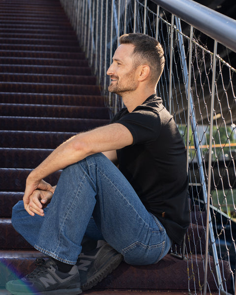 A side profile view of a smiling man sitting on outdoor, rusted steel stairs next to a metal wire railing. He is wearing a black short-sleeved v-neck t-shirt, blue jeans, and dark sneakers, resting his arms comfortably on his knees while looking forward.