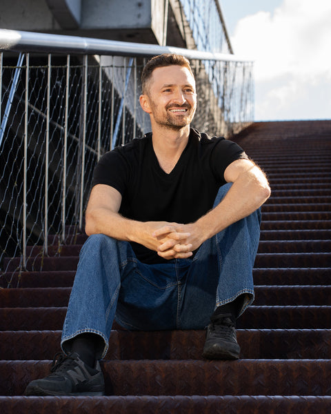 A smiling man sits on a wide set of outdoor, rusted steel stairs. He is wearing a black short-sleeved v-neck t-shirt, blue jeans, and dark sneakers, with his hands clasped together in his lap, looking off to the right.