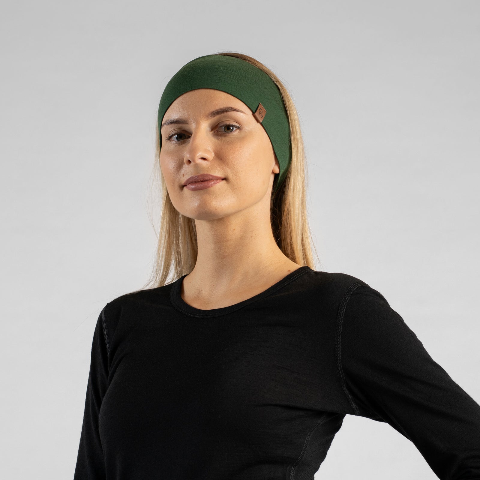 Billedet viser en kvinde, der sidder i en skov iført det unisex mørkegrønne menique merino PANDEBÅND. Hun har også en ærmeløs sort merino top på. Hun kigger til siden.