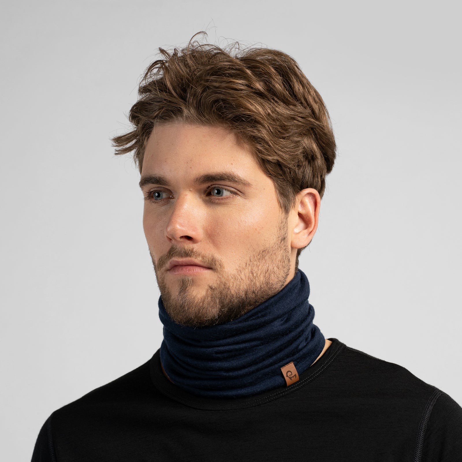 A dark navy blue tubular neck gaiter displayed against a solid white background. The soft fabric is styled with natural-looking folds and slouching, and a small, rectangular brown leather logo tag is attached near the bottom edge.