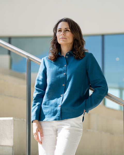 En kvinde med brunt hår, iført en koboltblå skjorte med knapper foran og hvide bukser med vide ben, står udendørs foran en moderne betonbygning.