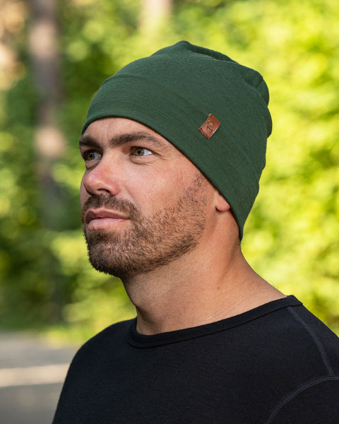A headshot of a man wearing the dark green merino beanie. It has a small menique logo on the side. His whole head is covered under the beanie. Behind him are trees in a blurred vision.