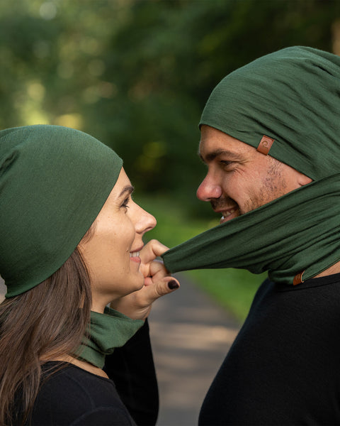 The picture features a man and a woman wearing the unisex dark green merino gaiter and beanie set. They are in a forest. The woman is pulling the man's gaiter to remove it. Both of them are smiling. 