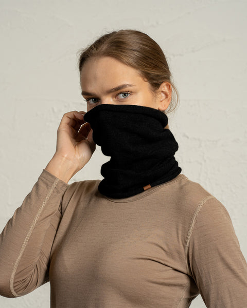 En kvinde iført en beige bluse med lange ærmer holder en sort uld snood-halsedisse over den nederste del af ansigtet, så mund og næse er dækket. Hendes blå øjne ser direkte ind i kameraet, og hendes hår er trukket tilbage i en pæn, lav frisure. Den strukturerede, lyse baggrund holder fokus på halsedissen og hendes udtryk.