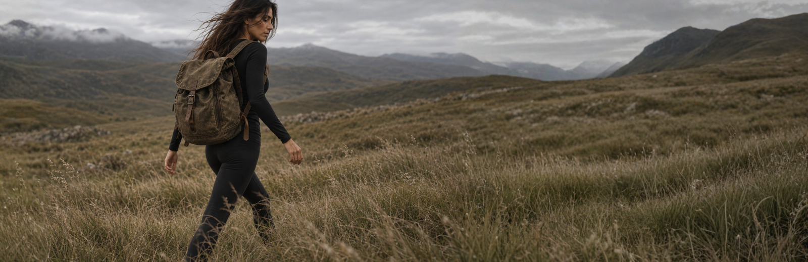 A woman with long dark hair walks through a grassy landscape while glancing back over her shoulder. She is dressed in a black long-sleeved top and leggings, carrying a weathered canvas backpack under an overcast sky.