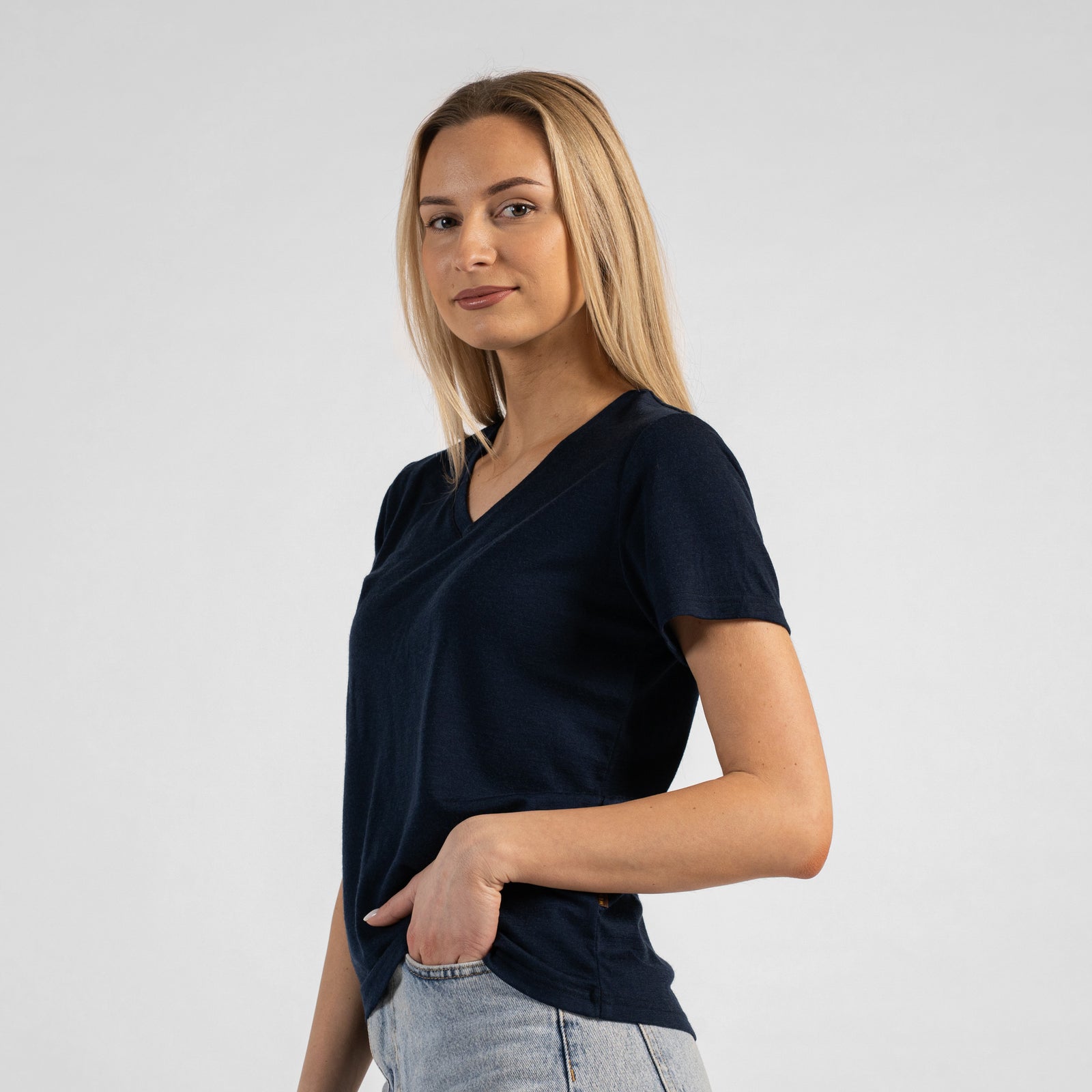 A studio portrait of a blonde woman leaning forward with a big smile, looking directly at the camera. Her hands are resting on her thighs just above her knees. She is wearing a regular fit, dark navy blue short-sleeved v-neck t-shirt and light-wash blue jeans against a plain light grey background.