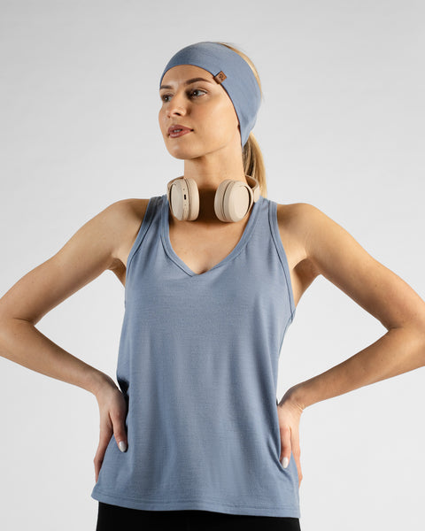 A studio portrait of a blonde woman standing with her hands on her hips, looking off to the left against a plain light grey background. She is wearing a dusty blue sleeveless sport top, a matching wide headband, black bottoms, and beige headphones resting around her neck.