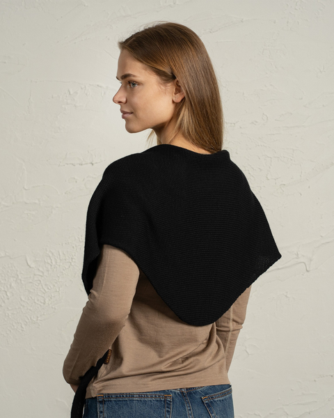 En kvinde, der står sideværts, bærer et sort strikket merinoulds-bandana, der er lagt over hendes skuldre og øvre ryg. Hun har glat, lysebrunt hår og er iført en beige bluse med lange ærmer og jeans, med en tekstureret lys væg i baggrunden.