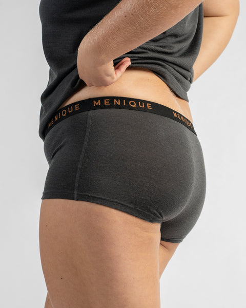 Side-back view of a model wearing dark grey boy-short underwear with a black elastic waistband featuring text, lifting the hem of a dark grey t-shirt.
