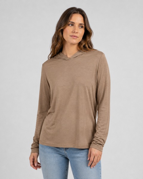 A studio portrait of a woman standing and looking off to her left against a plain light grey background. She has long wavy brown hair and is wearing a lightweight beige long-sleeved hoodie with the hood down, paired with light wash denim jeans.