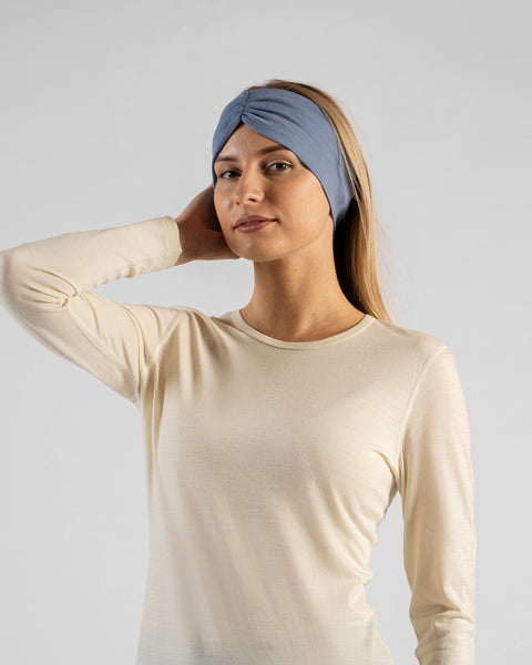 A studio portrait of a blonde woman wearing a light beige long-sleeved shirt and a wide dusty blue headband with a gathered twist detail at the front, looking directly at the camera with one hand resting behind her neck against a plain light grey background.