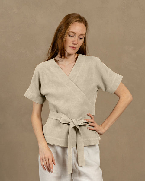 En lys hudfarvet person med langt, kastanjebrunt hår, iført en naturlig V-hals hørviklebluse med korte, vide ærmer og lyse bukser. De holder enderne af blusens bindebånd foran sig og danner en sløjfe.