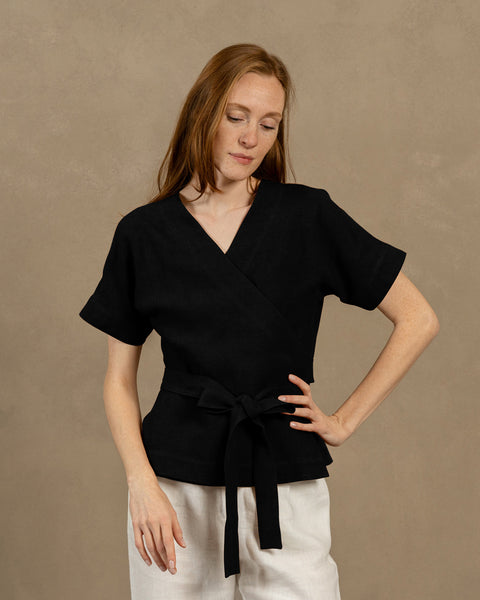 En lys hudfarvet person med langt, kastanjebrunt hår, iført en sort V-hals hørviklebluse med korte, vide ærmer og lyse bukser. De holder enderne af blusens bindebånd foran sig og danner en sløjfe.