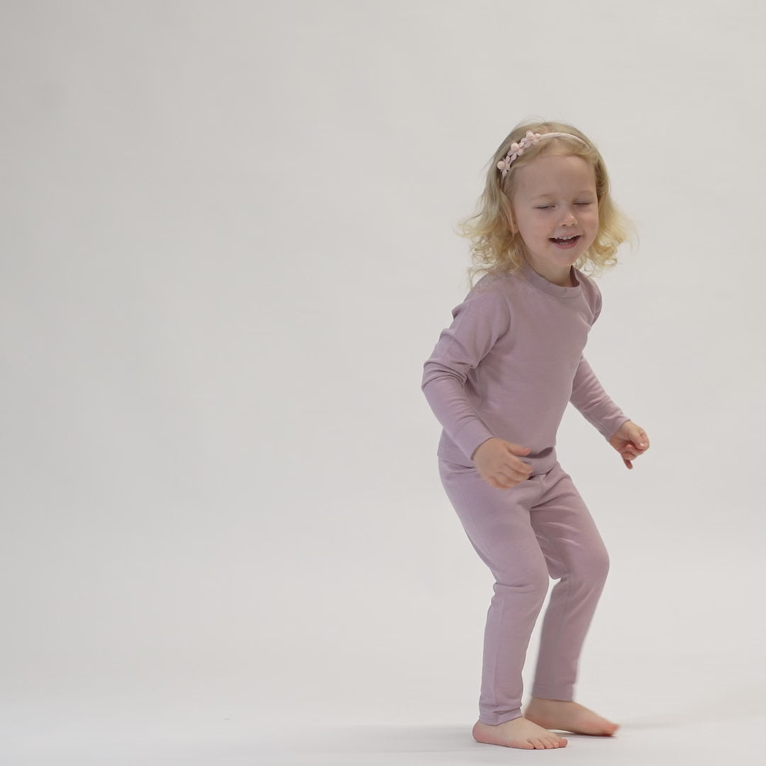 A child with blonde, curly hair and a pink floral headband is captured mid-movement against a white background. The child is wearing a dusty pink long-sleeved top and matching pants, and is smiling with eyes closed, appearing to be jumping or dancing.
