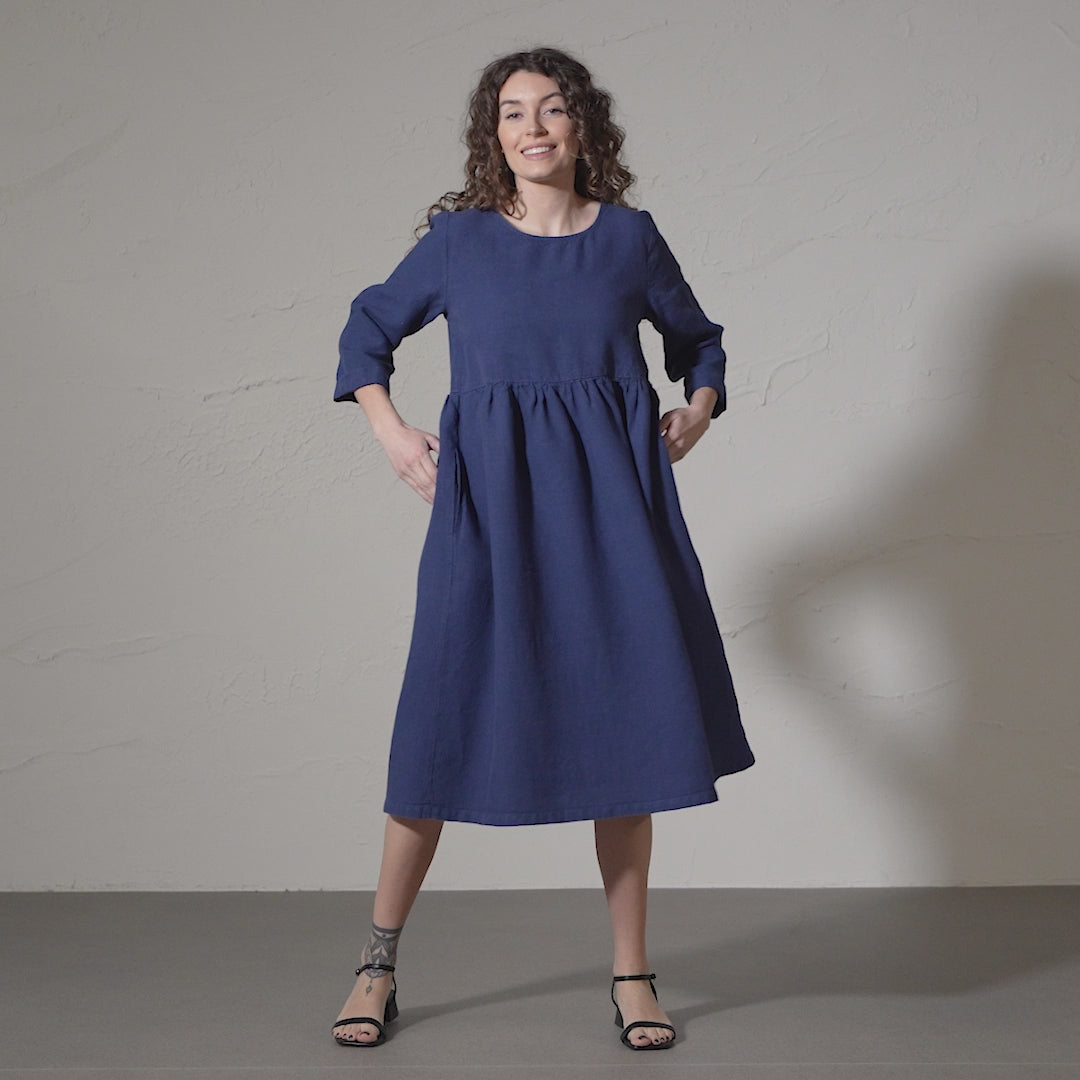 A woman with curly brown hair smiles and models a long-sleeved, storm blue linen dress with a gathered waist and pockets against a textured gray backdrop in a short video. 