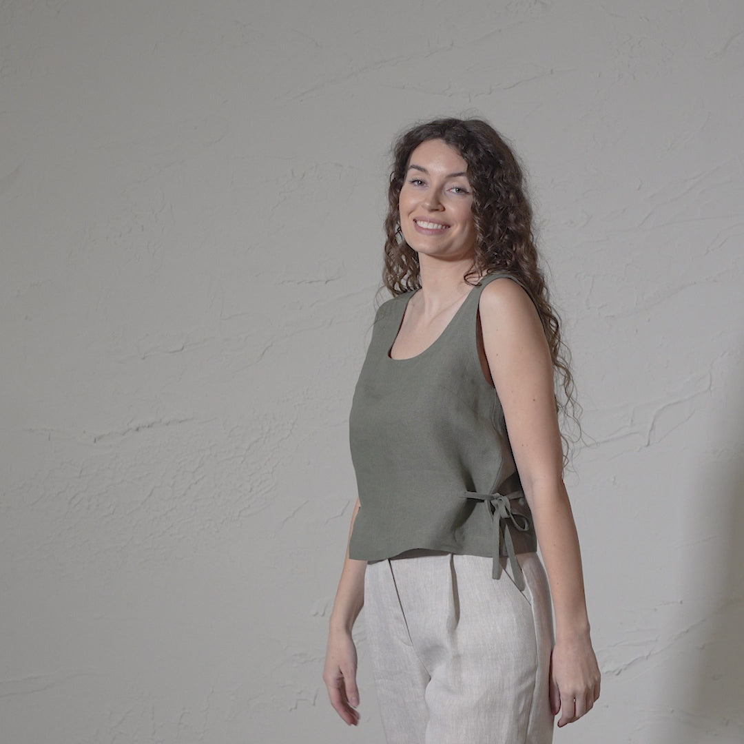 A smiling woman with long, curly brown hair models a sleeveless stone green linen top with a side tie and menique linen pants in a short video against a textured off-white wall.