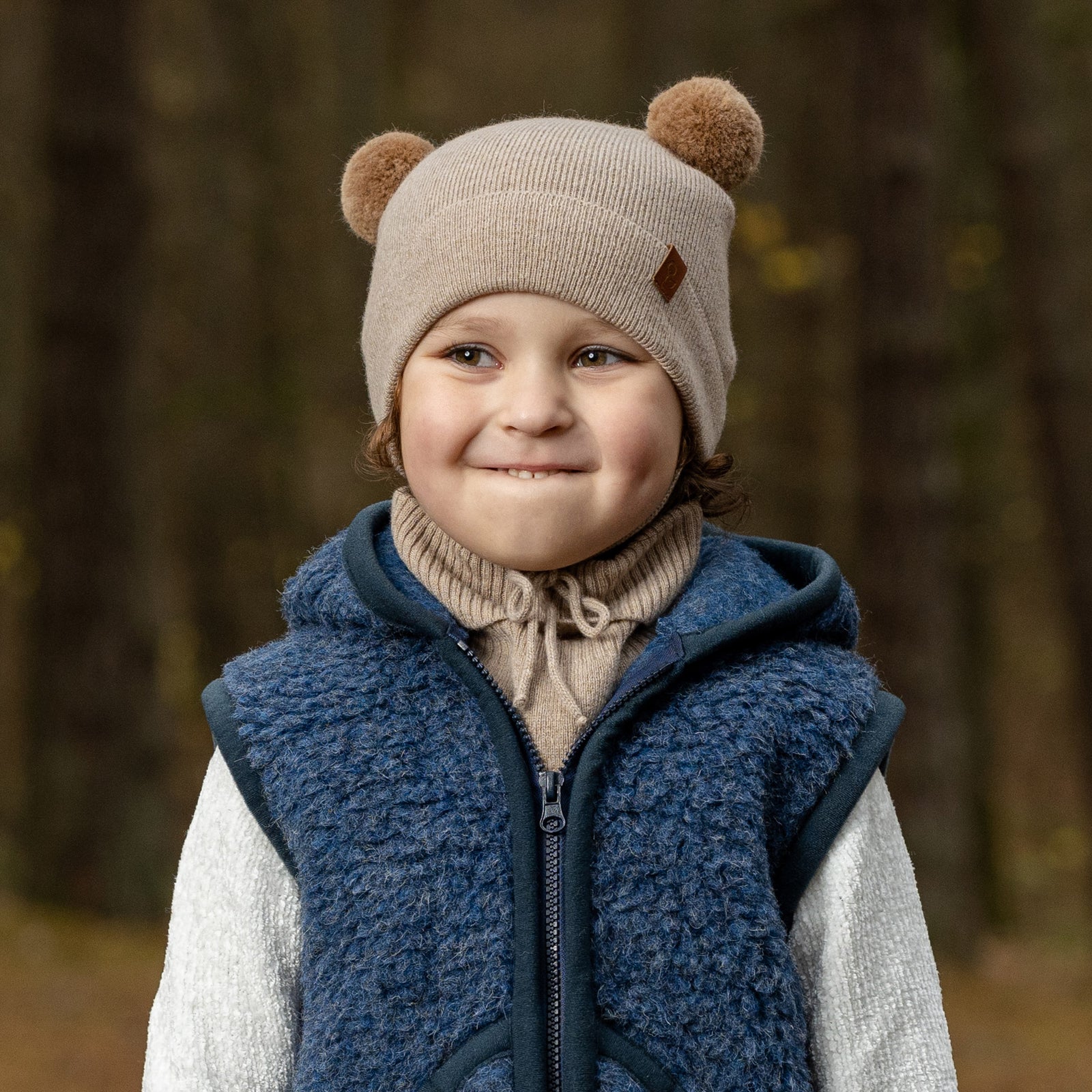 A young child with blue eyes and a wide smile, is looking directly at the viewer while peeking over a metal playground railing. They are wearing a beige knit hat with brown pom-pom ears, beige mittens, and a light pink sherpa-style coat.